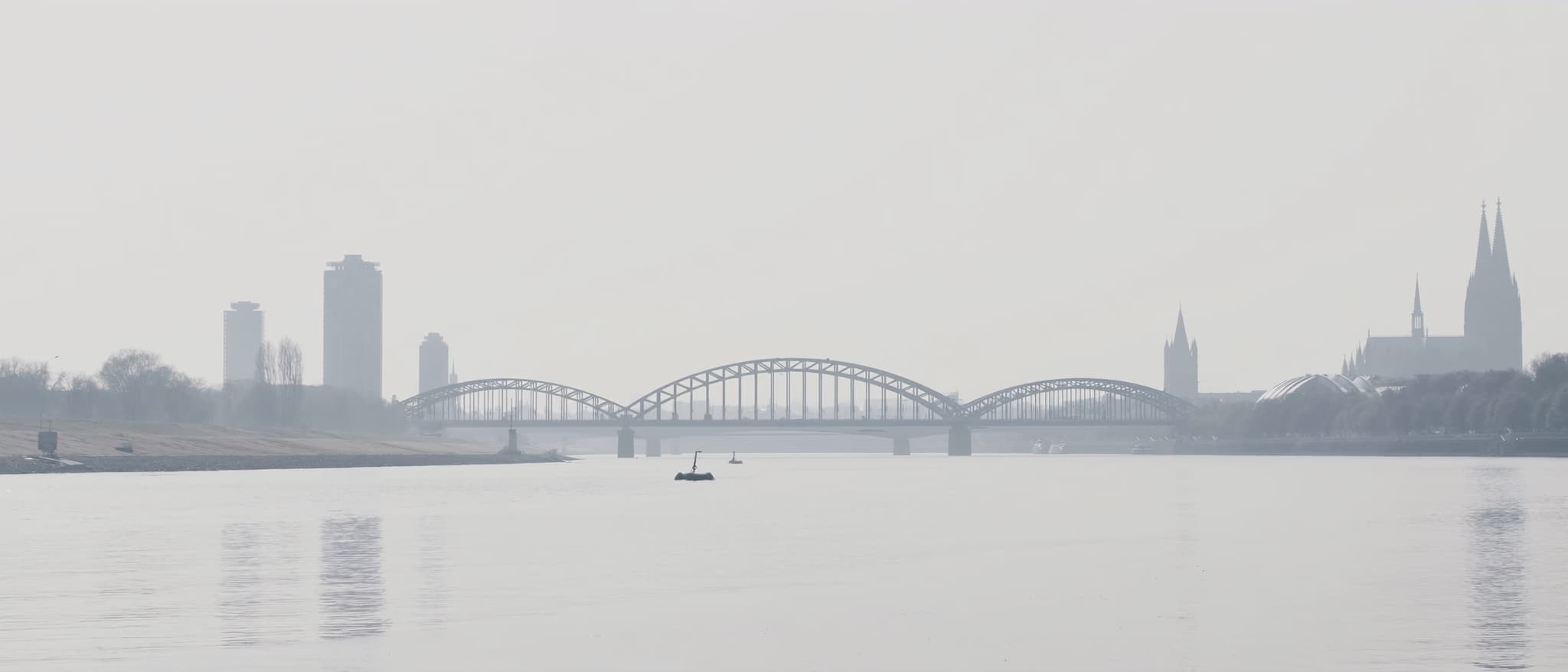 Eine Stahlfachwerkbrücke über den Rhein im Dunst, dahinter die Türme der Stadt als Schatten